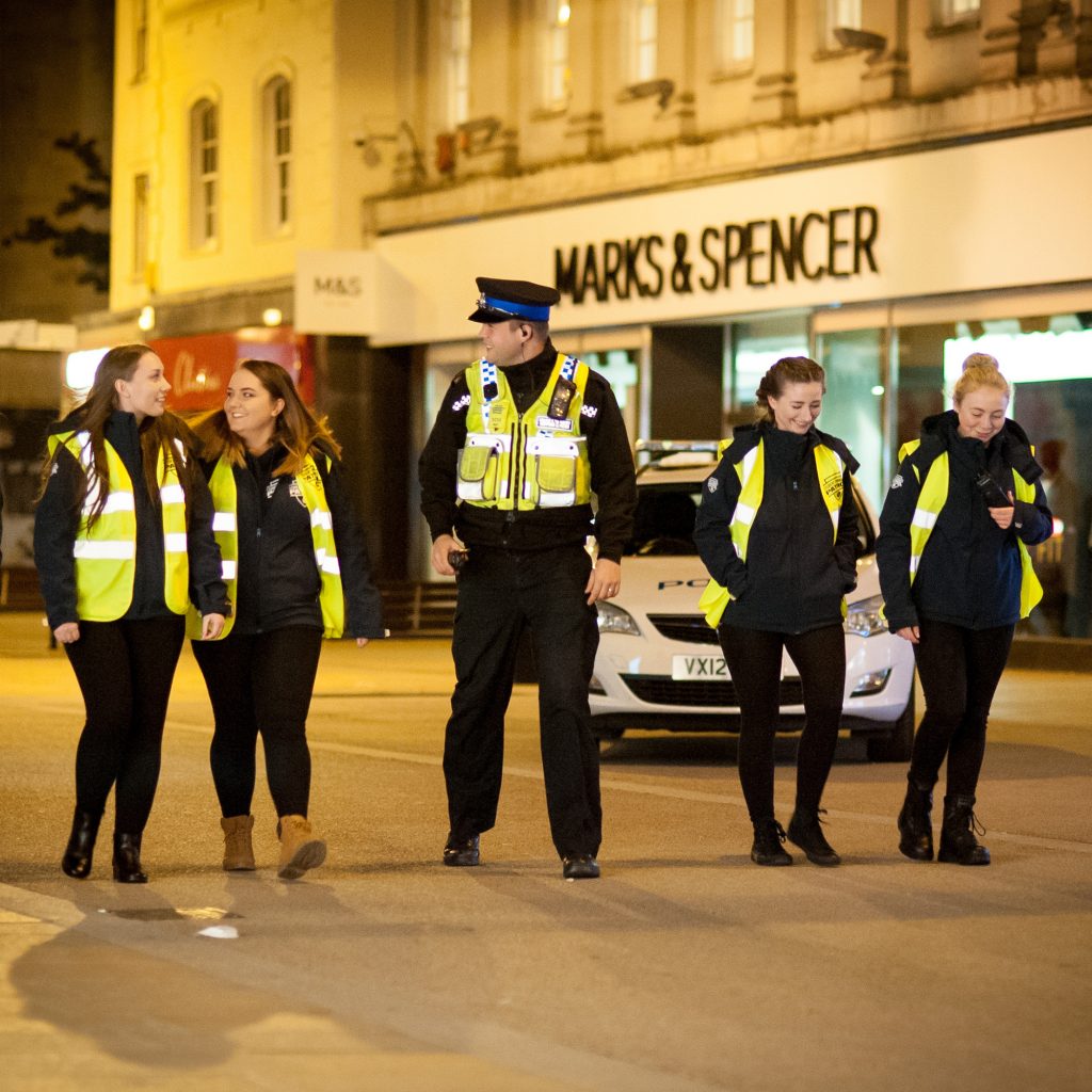 Community Police officer and students walking down Chelteham High Street at night in reflective clothing