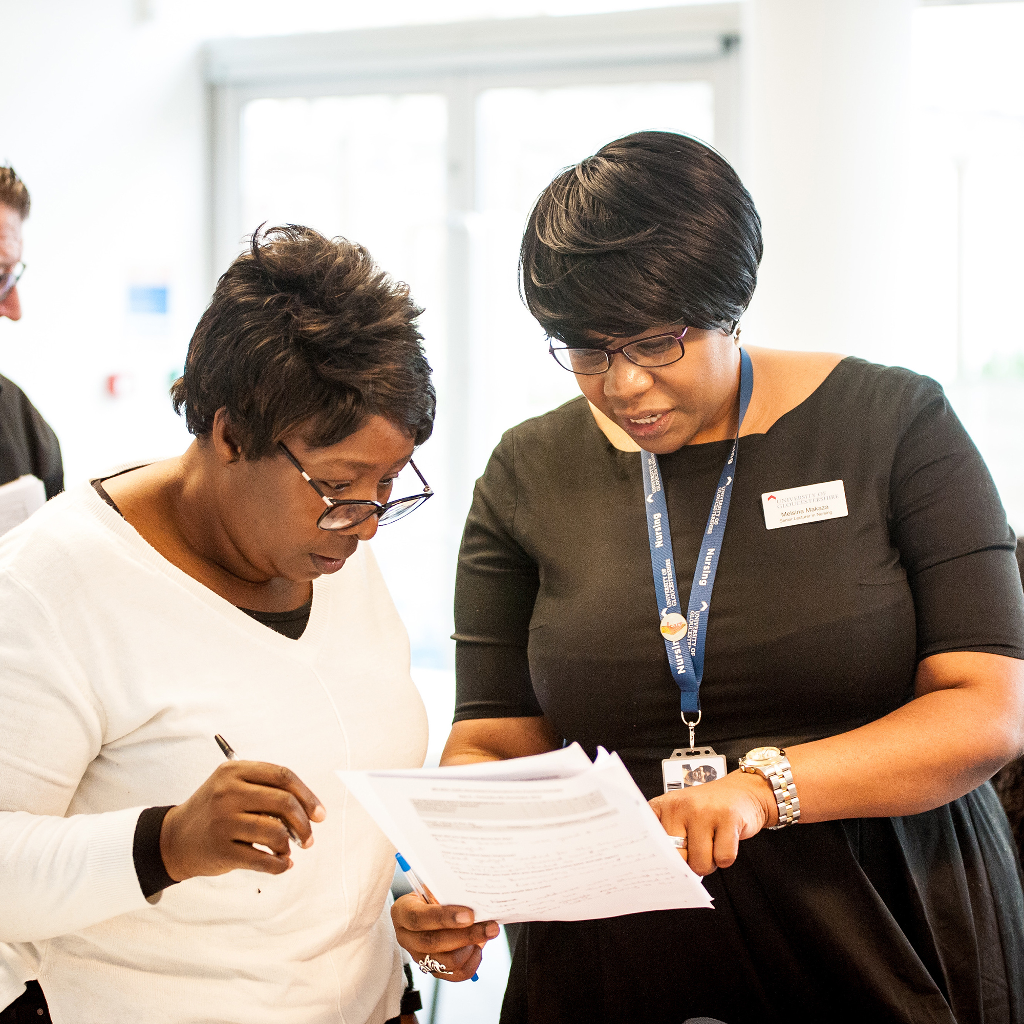 Woman in black dress pointing out something on a piece of paper to woman in white top