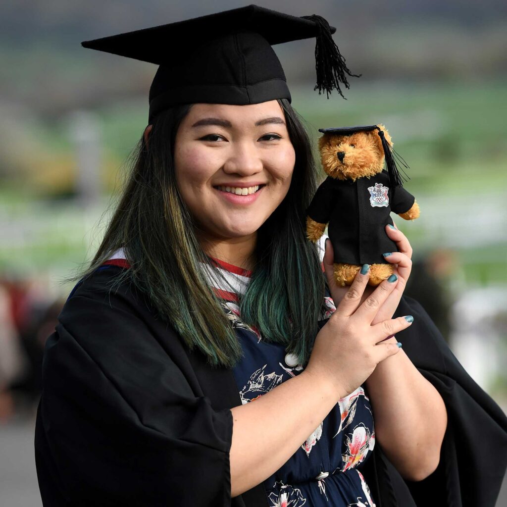 A student wearing graduation gown and hat holds 'Brad the Grad' little teddy bear.