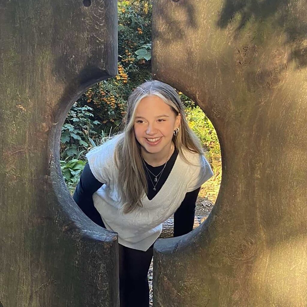 Student Ellie-may looking through a circular hole in a piece of carved wood in an outdoor setting.
