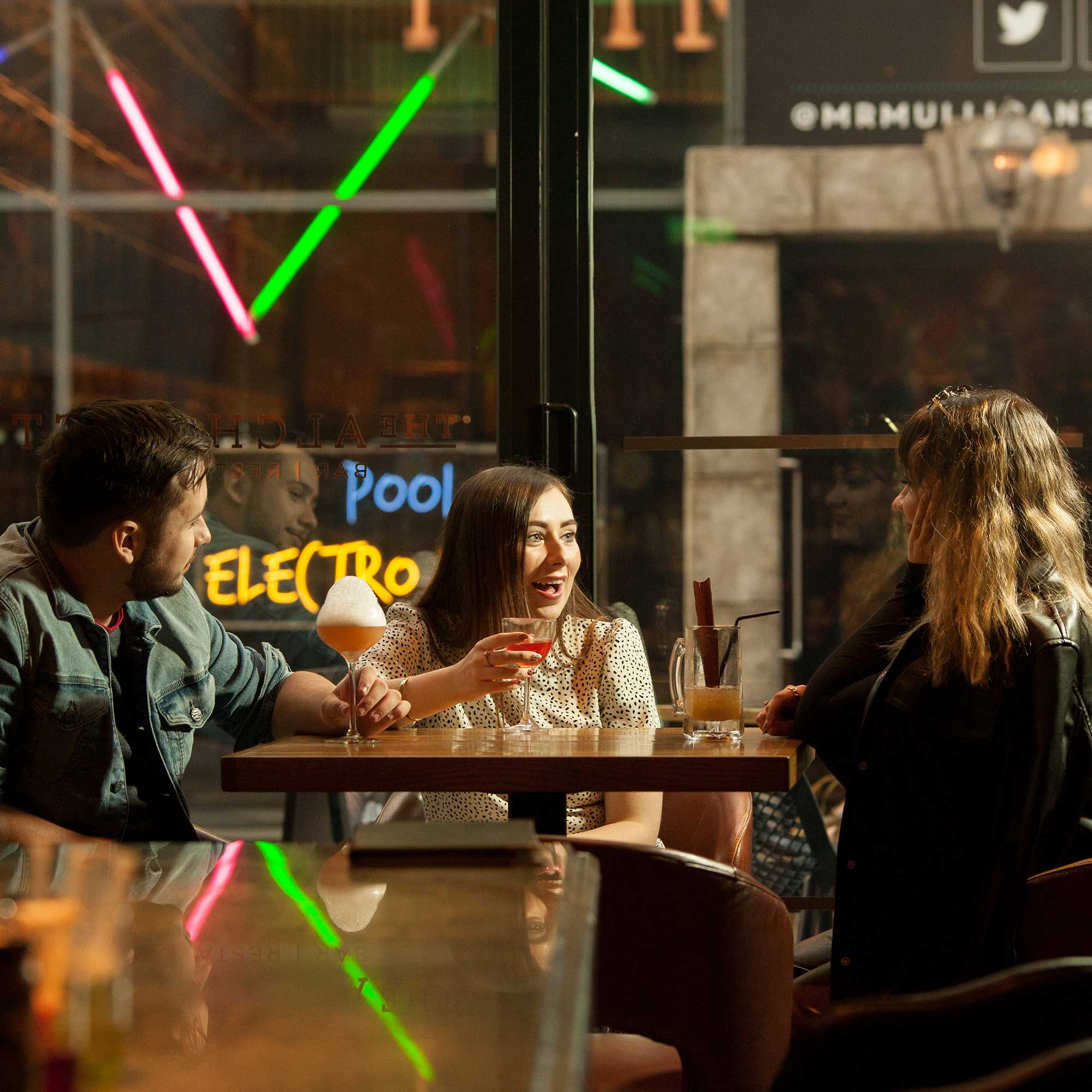 Three students sit at a table in a bar drinking cocktails.