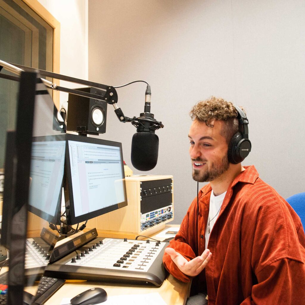 Student sitting in front of mixing desk in a radio studio. He's wearing headphones and talking into a microphone.