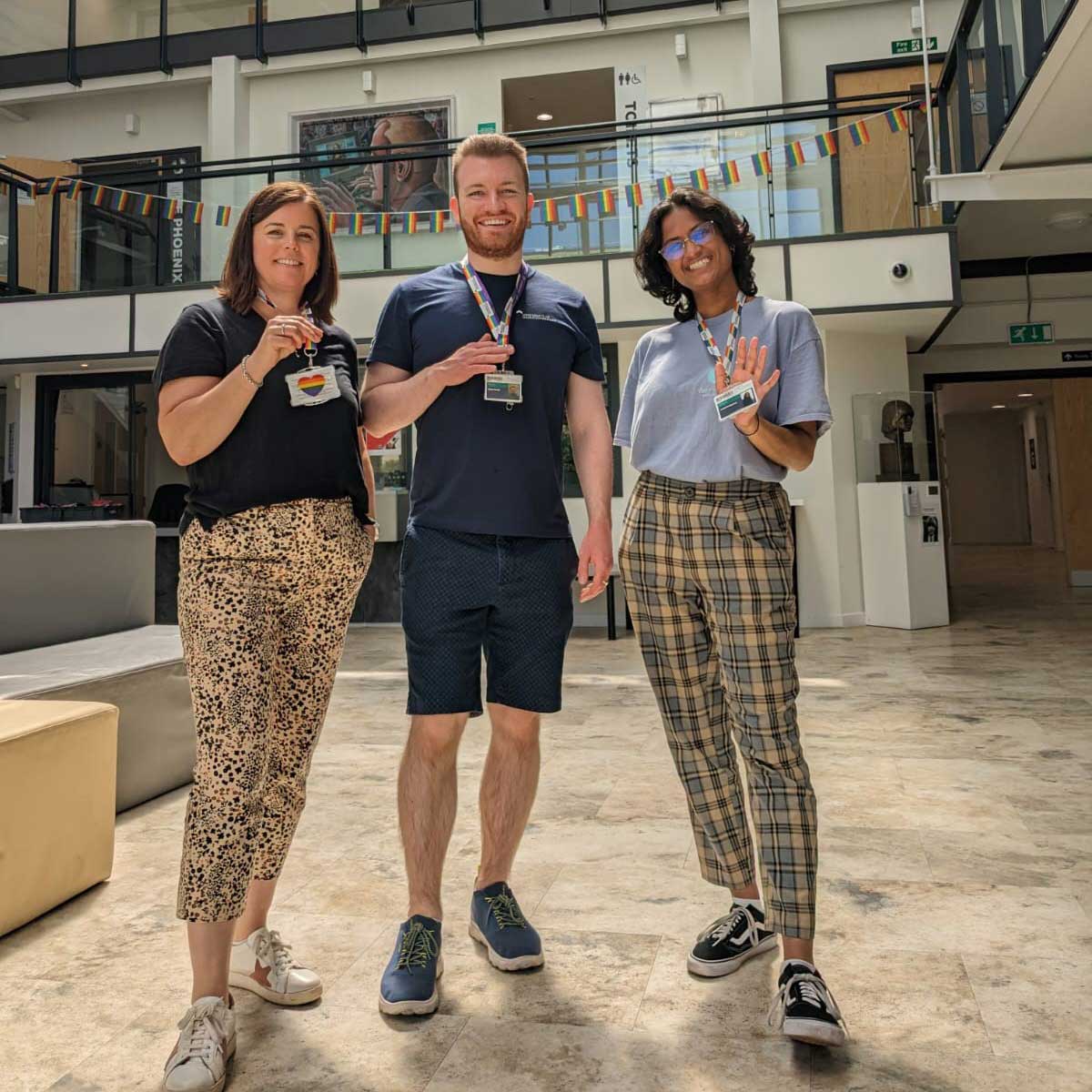 Three staff members standing in the atrium holding their rainbow lanyards and smiling.