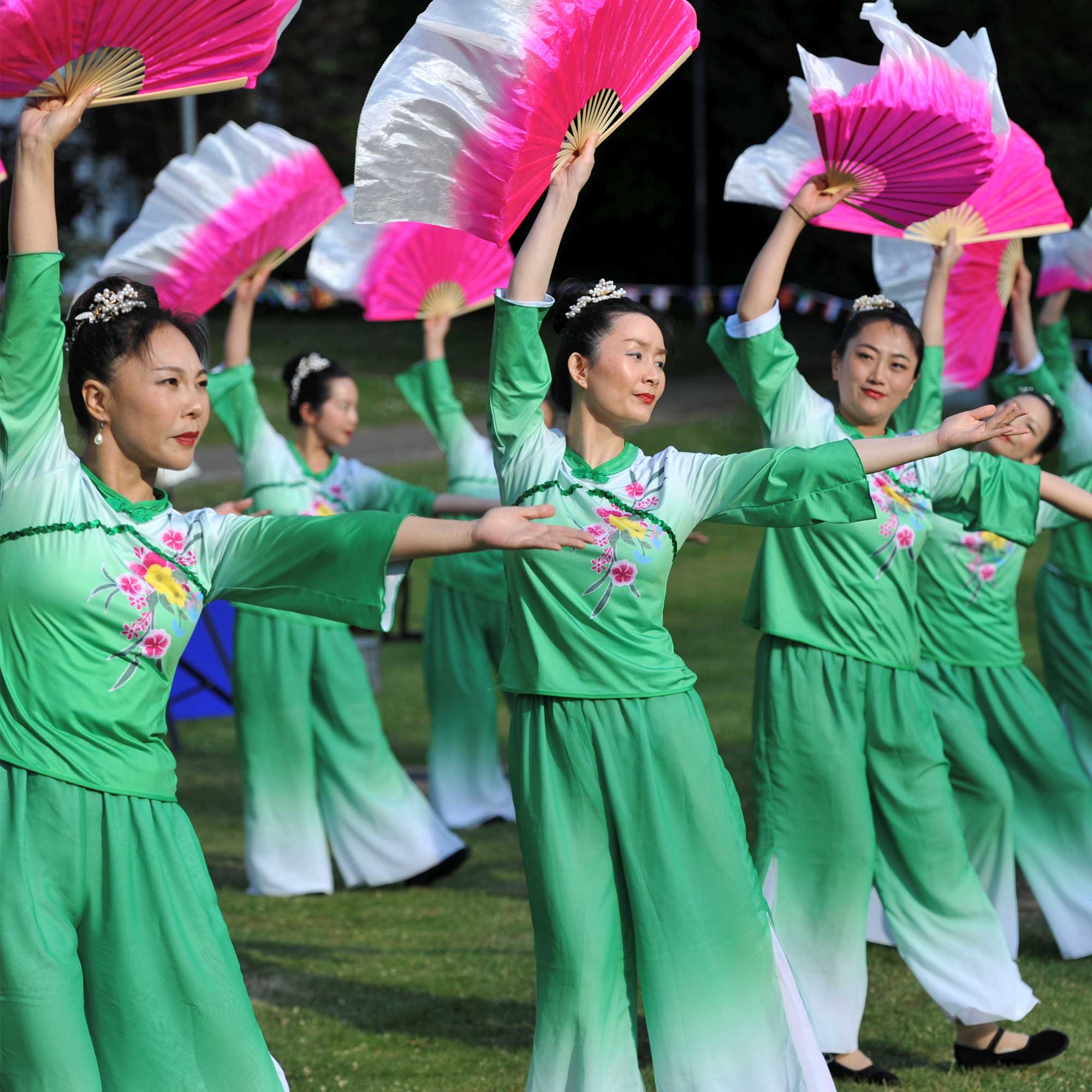 Female dancers wearing green outfits holding pink fans above their heads as they dance.