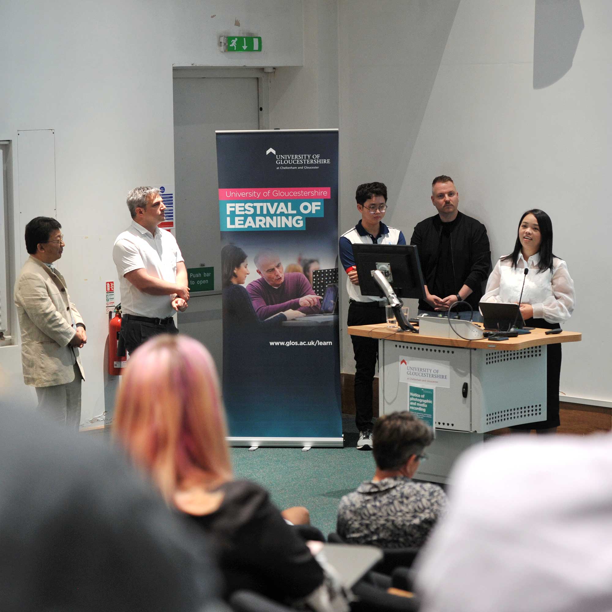 A speaker gives a talk at the front of a lecture theatre next to a banner displaying the text, Festival of Learning.