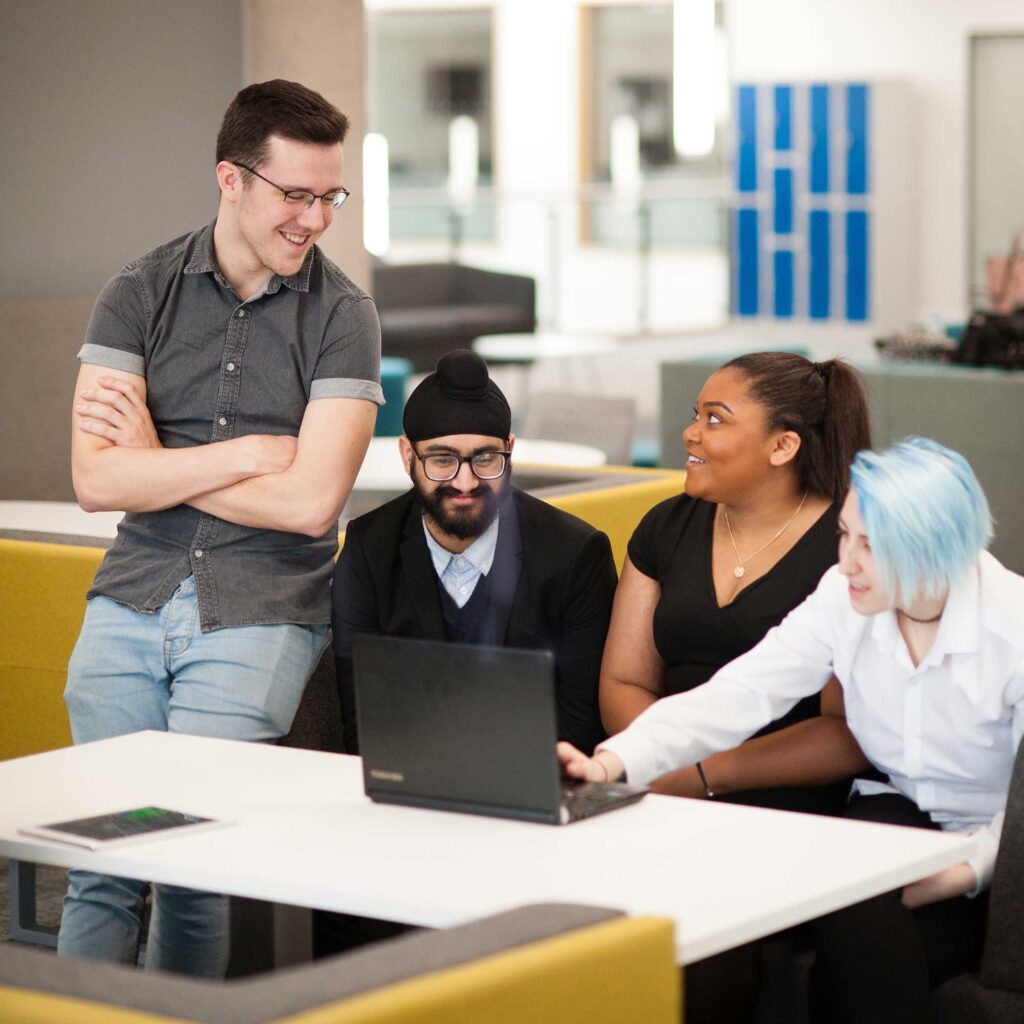 Four students sit at a desk talking while using a laptop.