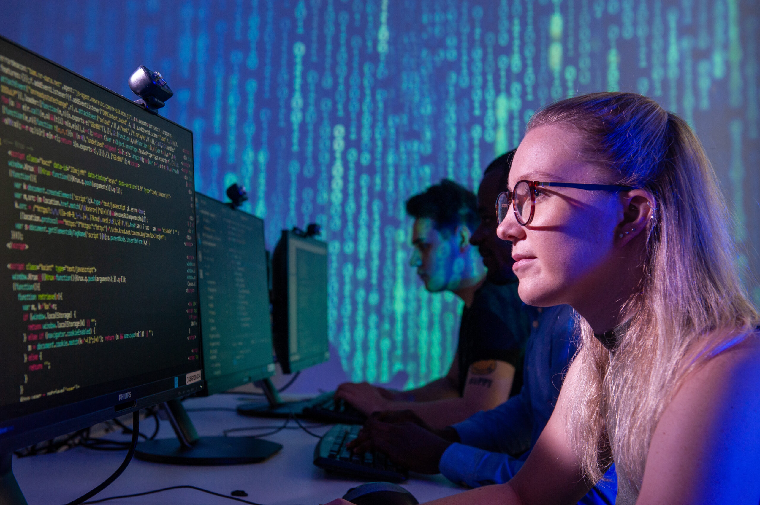 A student working on a computer in front of a screen showing strings of binary code.
