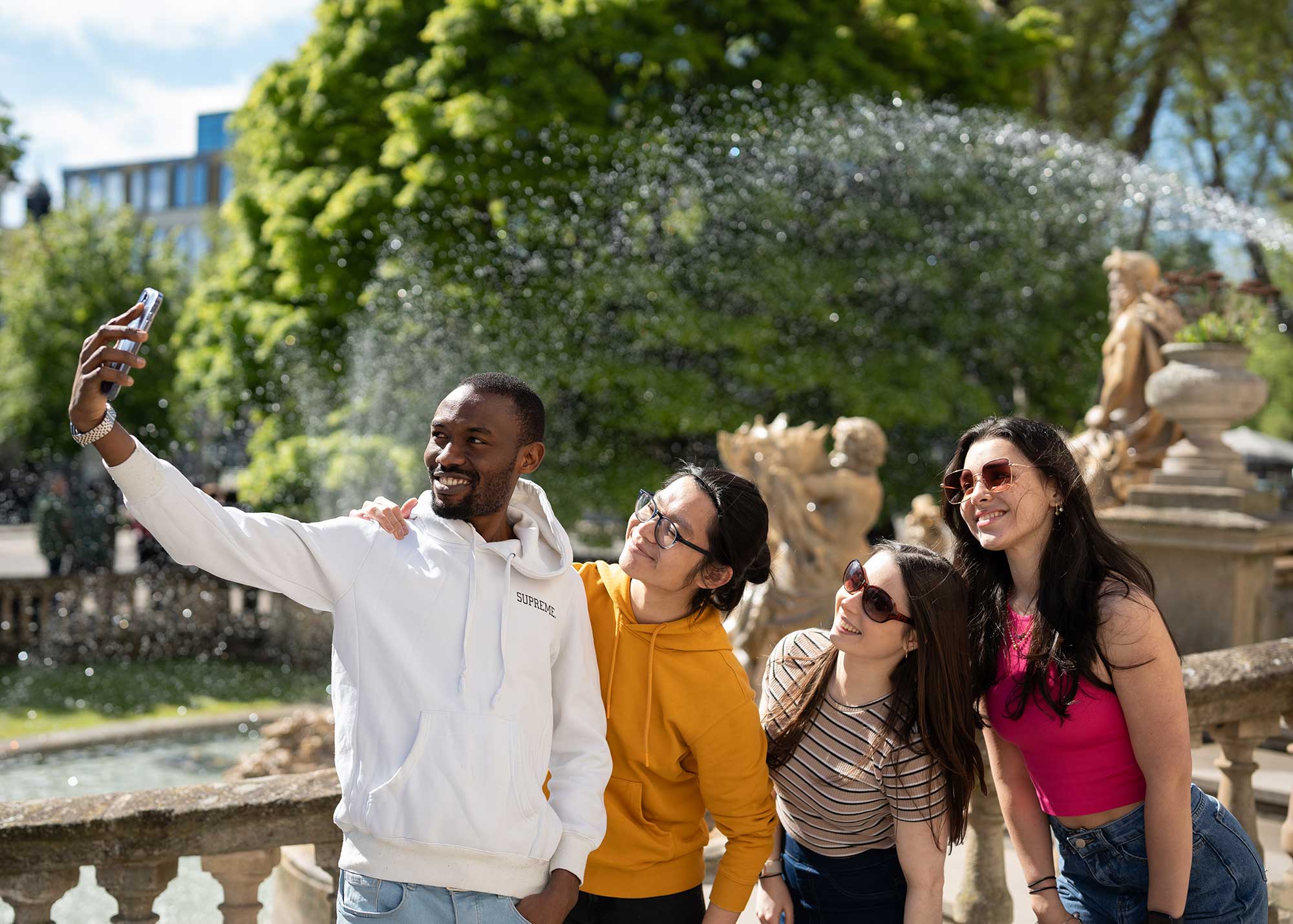A group of four students take a selfie in front of The Neptune Fountain in Cheltenham.