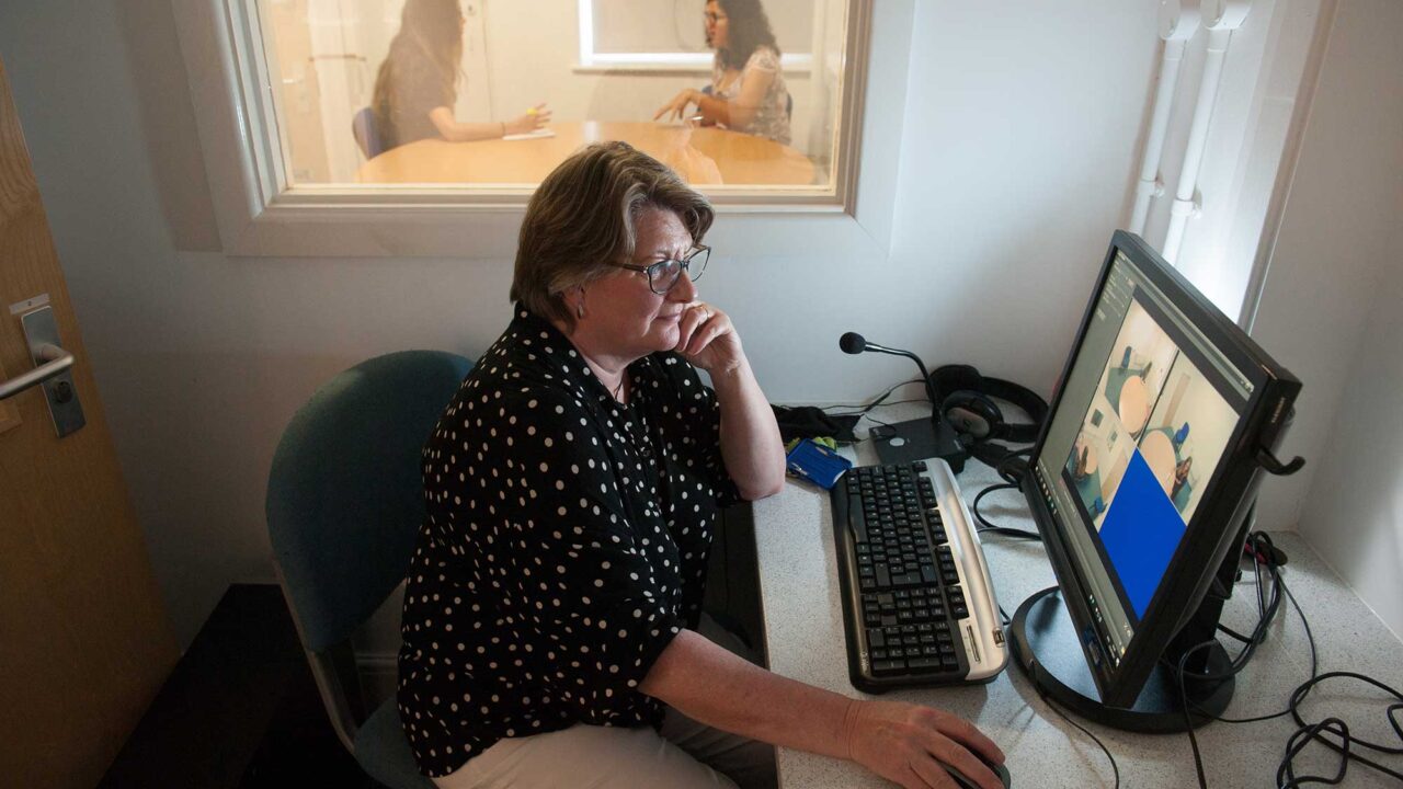 A woman looks at a screen with CCTV feeds while an interview takes place in the adjacent room.
