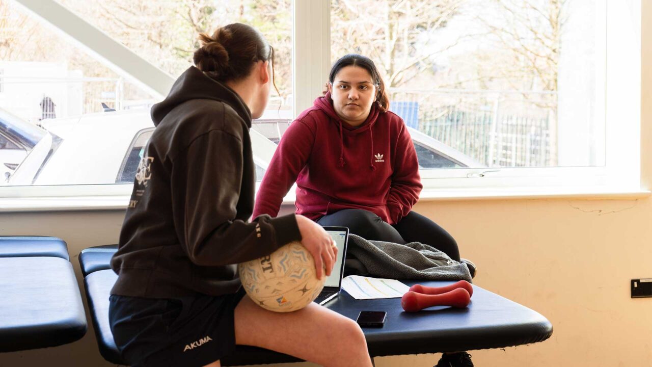 Two students in a sports therapy session, sat on a treatment bed with laptop, netball and small weights.