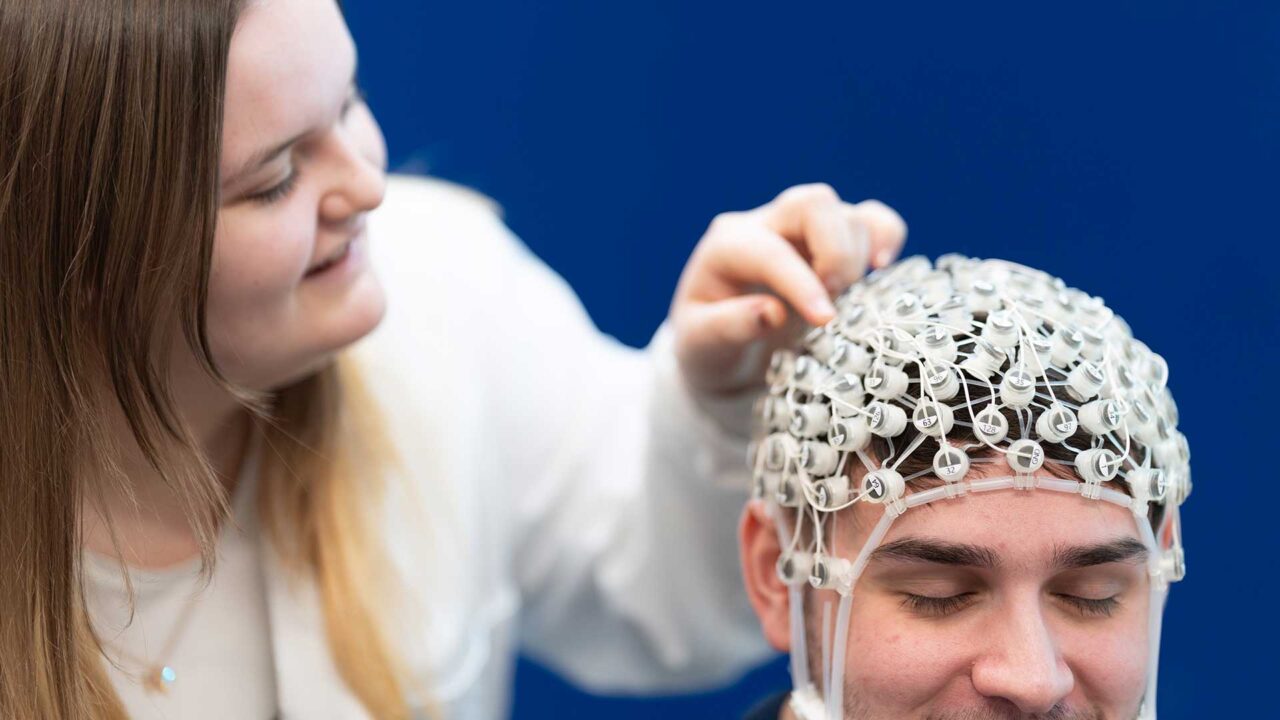 A student examines the EEG cap that a fellow student is wearing on their head.