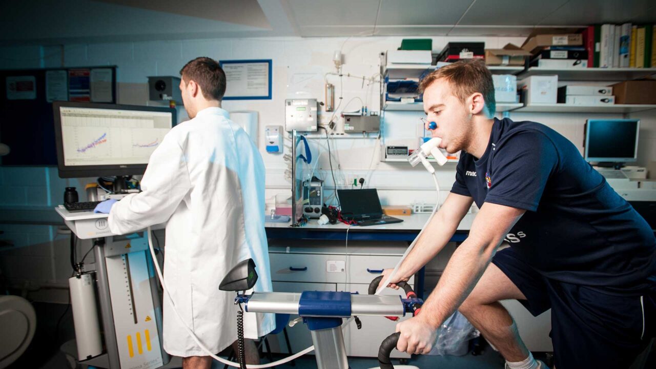 Student on an exercise bike undertaking a sports science test, student in white lab coat looking at results on computer screen in the foreground.